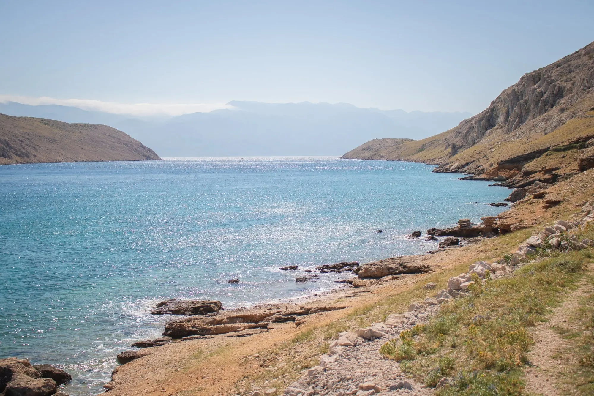 Felsige Bucht mit glitzerndem türkisem Meer, im Hintergrund Berge im Dunst