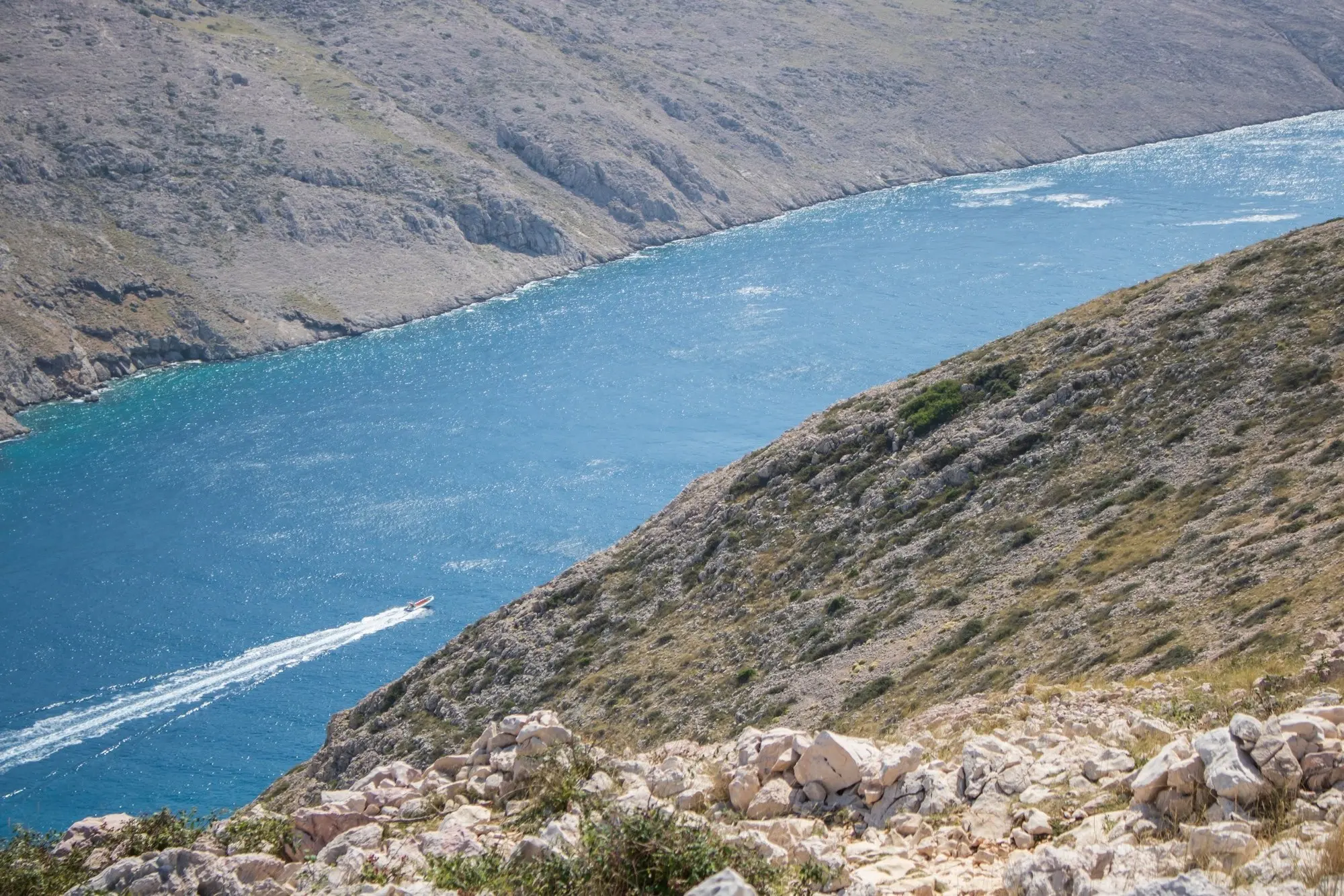 Blick von einem steinigen Hang auf eine Meerenge, ein Motorboot zieht eine weiße Spur